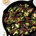 A skillet of Beef and Broccoli and small bowls of green onions and sesame seeds to the side.