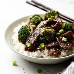 Beef and Broccoli Stir Fry in a bowl over rice with chop sticks on the side.