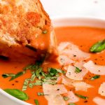 Dipping a grilled cheese sandwich into a bowl of tomato soup topped with basil and parmesan.