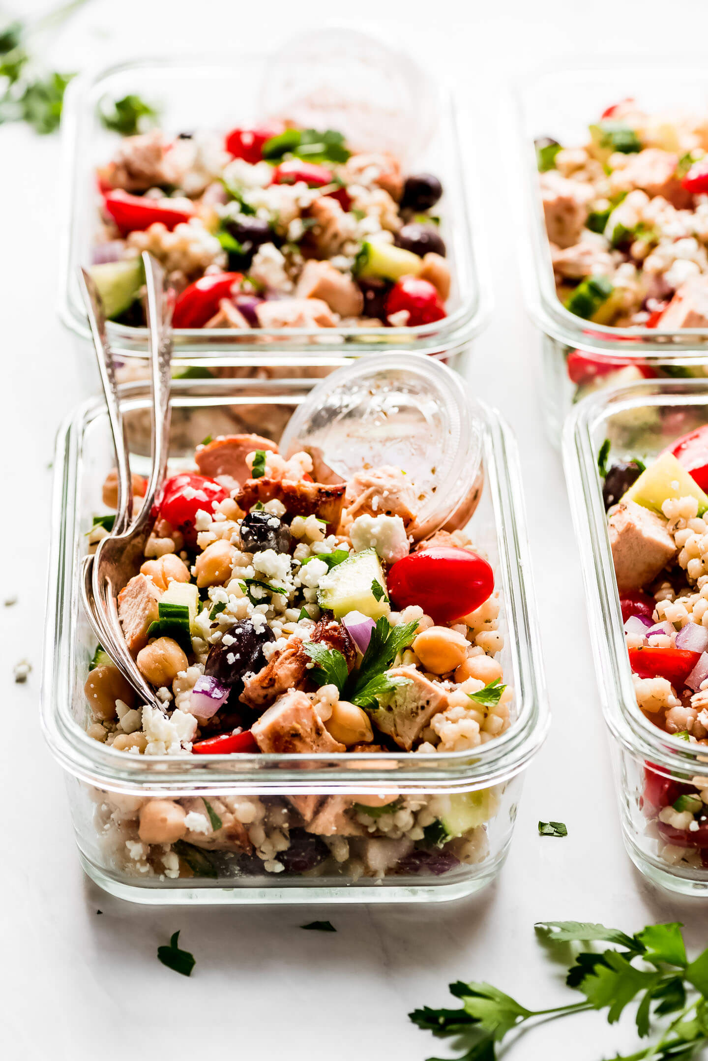 A meal prep container of Greek Couscous Salad with a small cup of dressing and two forks on the side.
