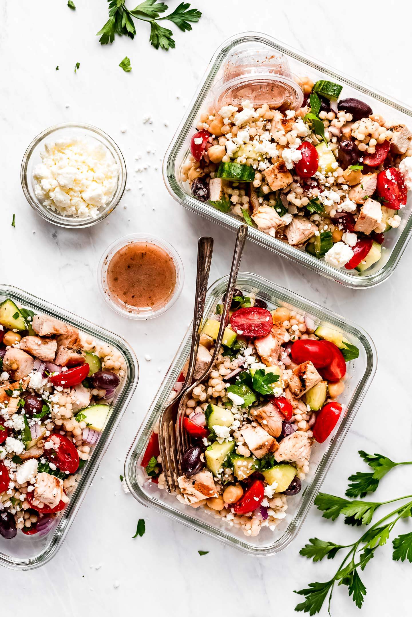 Three glass meal prep containers of Mediterranean Couscous Salad, small containers of dressing, a bowl of feta cheese.