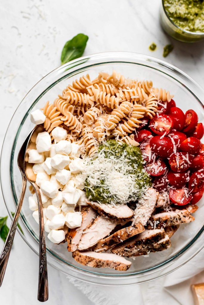 A large mixing bowl of rotini pasta, halved grape tomatoes, grilled chicken, mozzarella pearls, pesto sauce, and grated Parmesan.