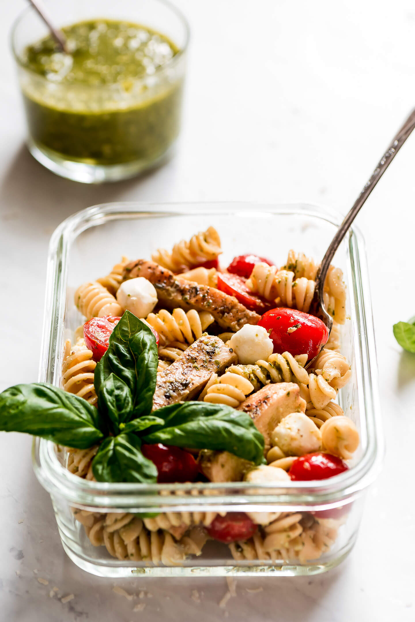 One glass meal prep container of Pesto Pasta Salad with a fork in the side and a bowl of pesto in the background.