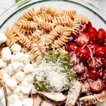 A large mixing bowl of rotini pasta, halved grape tomatoes, grilled chicken, mozzarella pearls, pesto sauce, and grated Parmesan.
