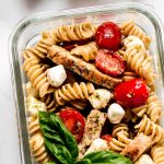 A glass meal prep container of rotini with grape tomatoes, chicken, mozzarella, and basil.