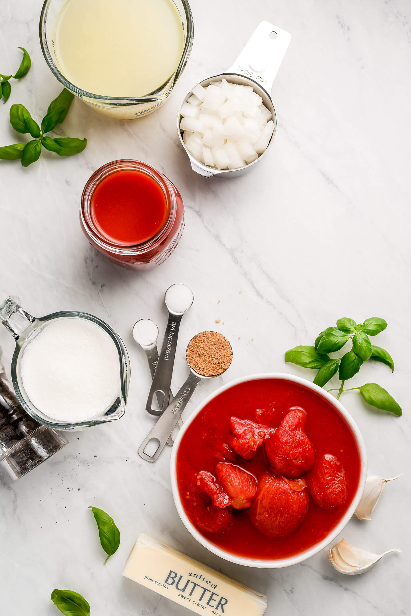 Ingredients on a marble surface- chicken broth, onions, tomato sauce, cream, whole peeled tomatoes, butter, garlic, and basil.