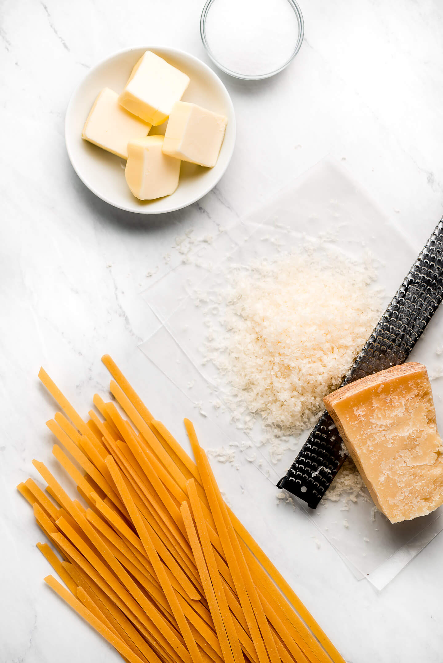 Ingredients on a marble surface: butter, salt, parmigiano reggiano, and dried fettuccine pasta.