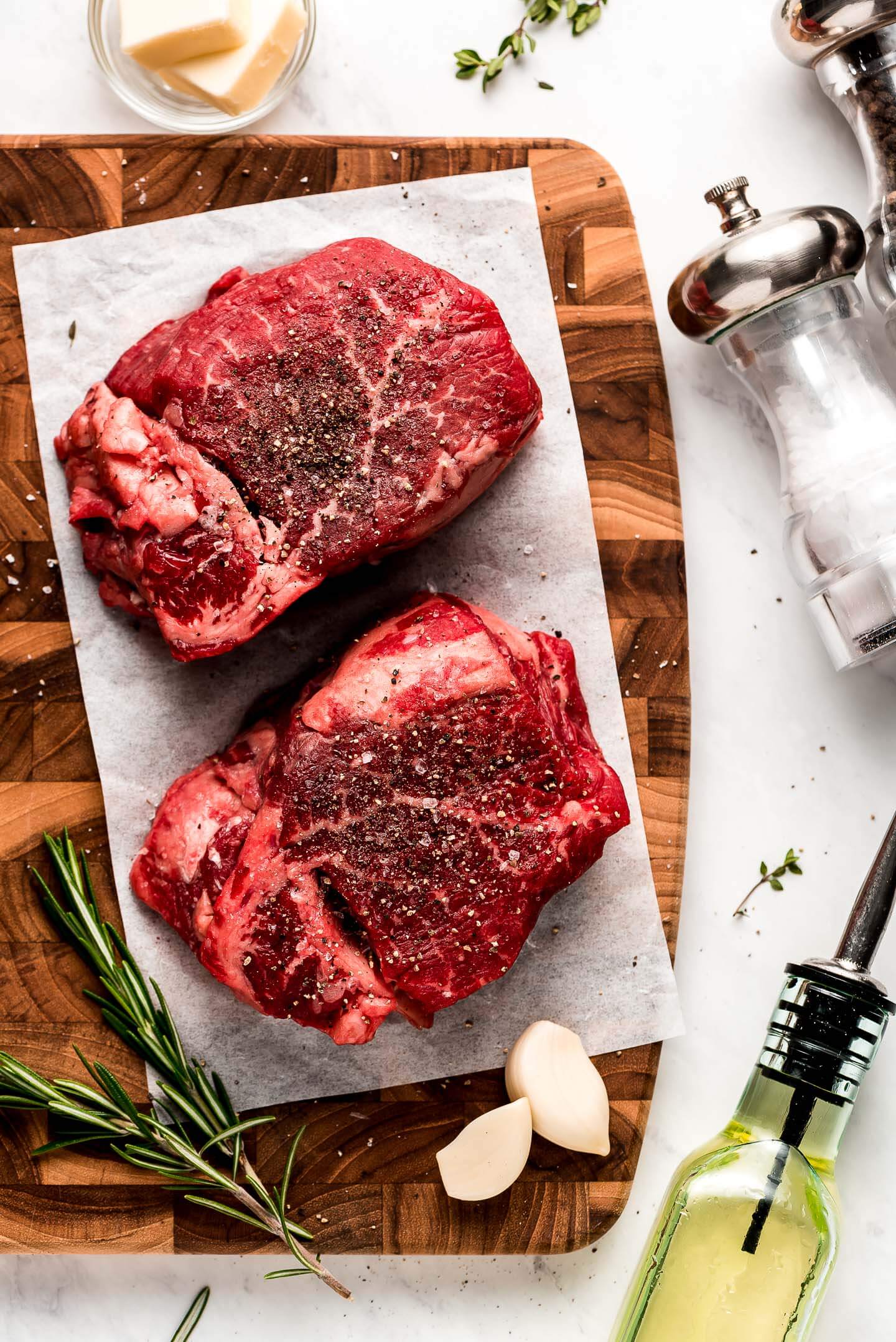 Two raw cuts of filet mignon on a wooden cutting board with fresh herbs, salt and pepper, olive oil, and butter on the table.