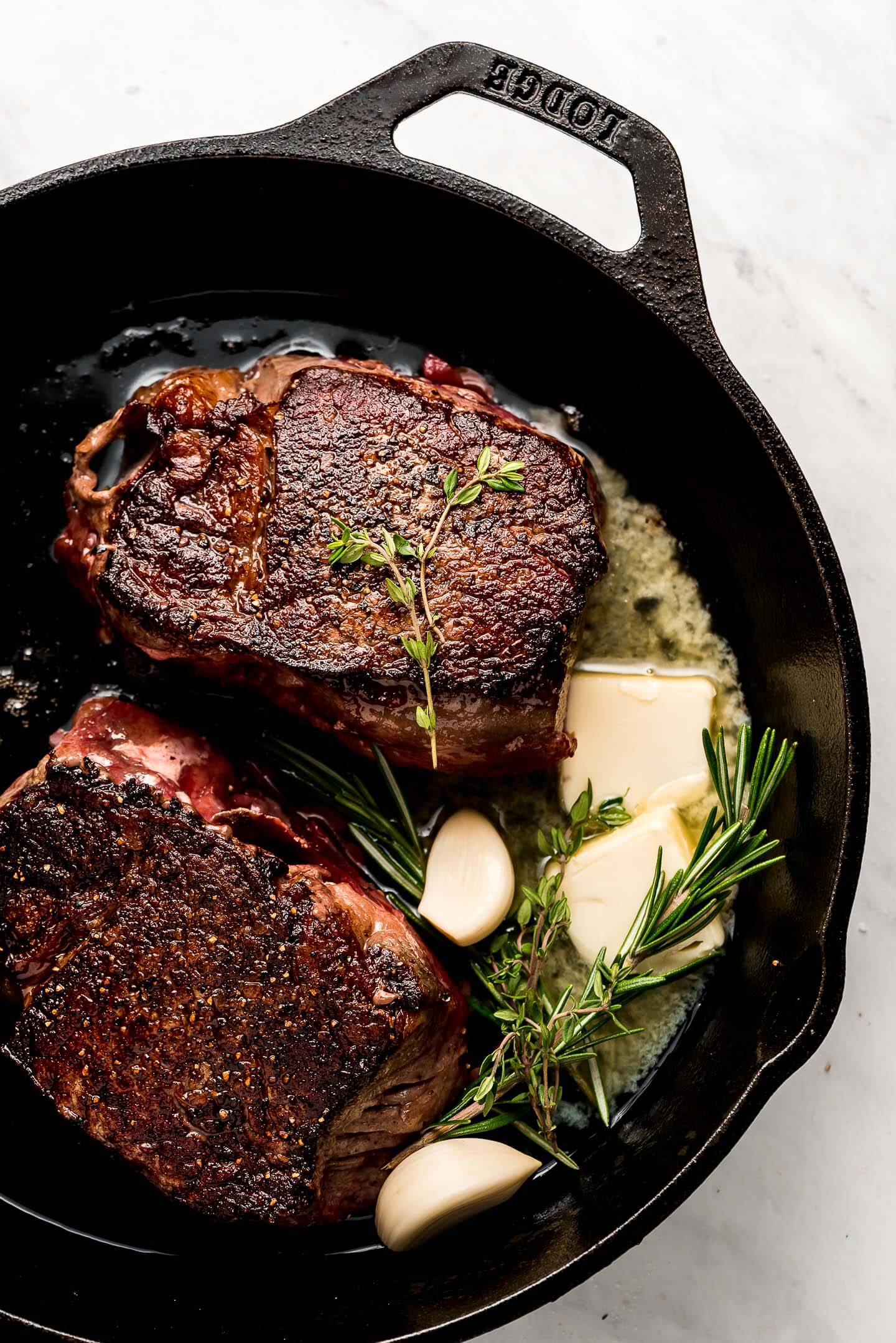 Two beef tenderloin steaks in a cast iron skillet with fresh herbs, butter, and garlic cloves.
