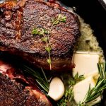 Two beef tenderloin steaks in a cast iron skillet with fresh herbs, butter, and garlic cloves.