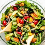 A large bowl of tossed salad comprised of romaine lettuce, olives, cucumbers, peppers, tomatoes, onions, croutons, sunflower seeds, and parmesan cheese.