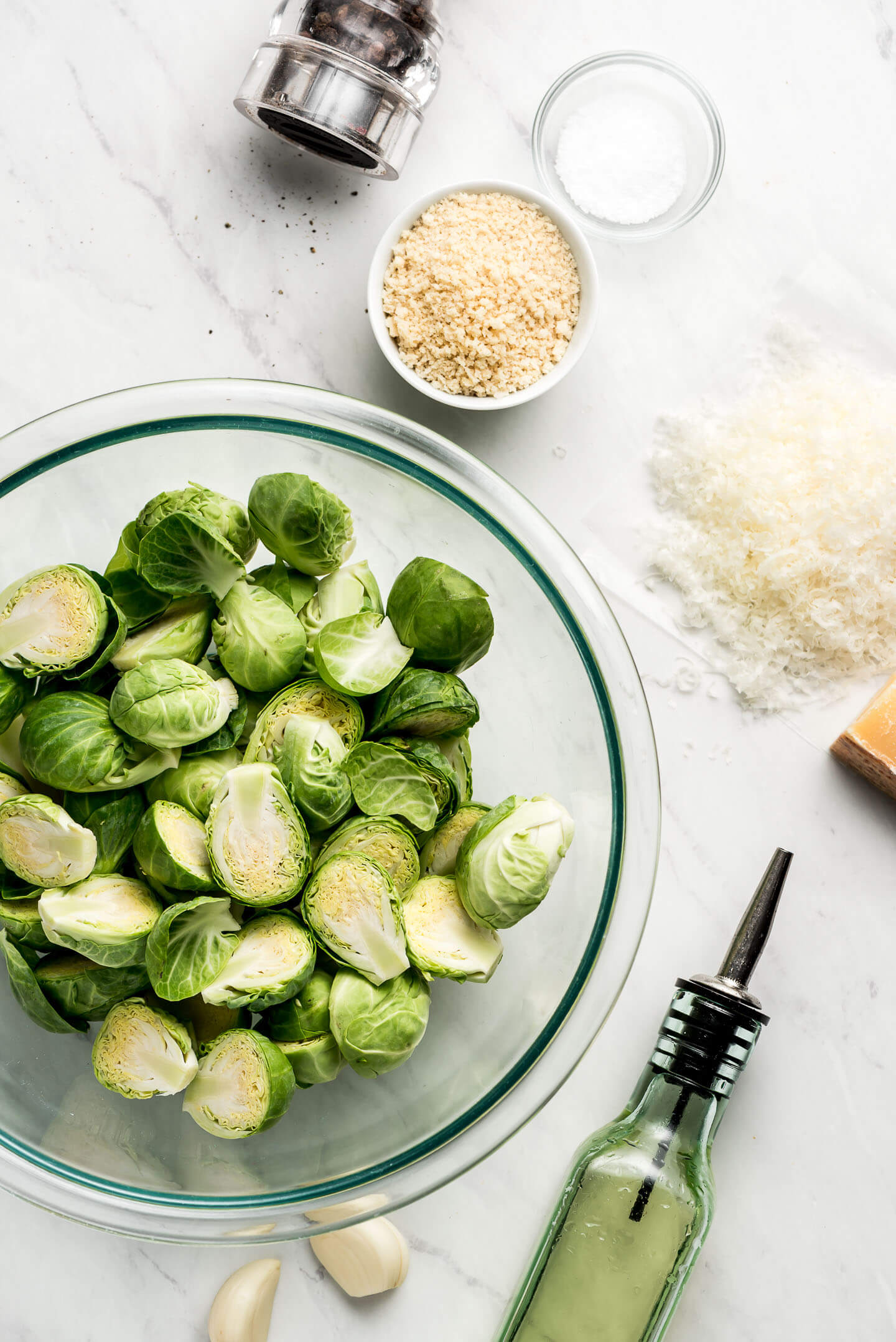 Halved Brussels Sprouts in a bowl, oil, garlic, grated parmesan, Panko, salt, and pepper on a marble surface.