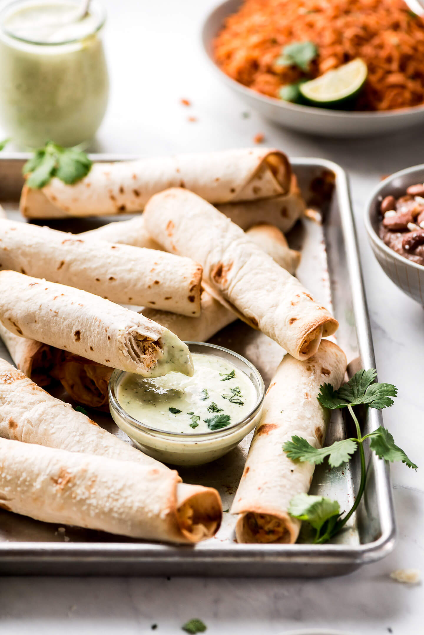 Many Chicken Taquitos on a baking sheet with dipping sauce and rice and beans in bowls in the background.