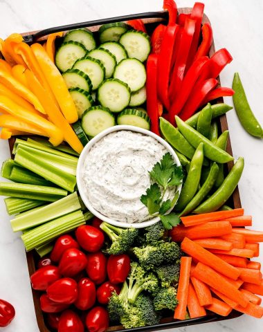 A large platter with a bowl Veggie Dip and sliced fresh vegetables- bell peppers, cucumbers, snap peas, carrots, broccoli, celery, and grape tomatoes.