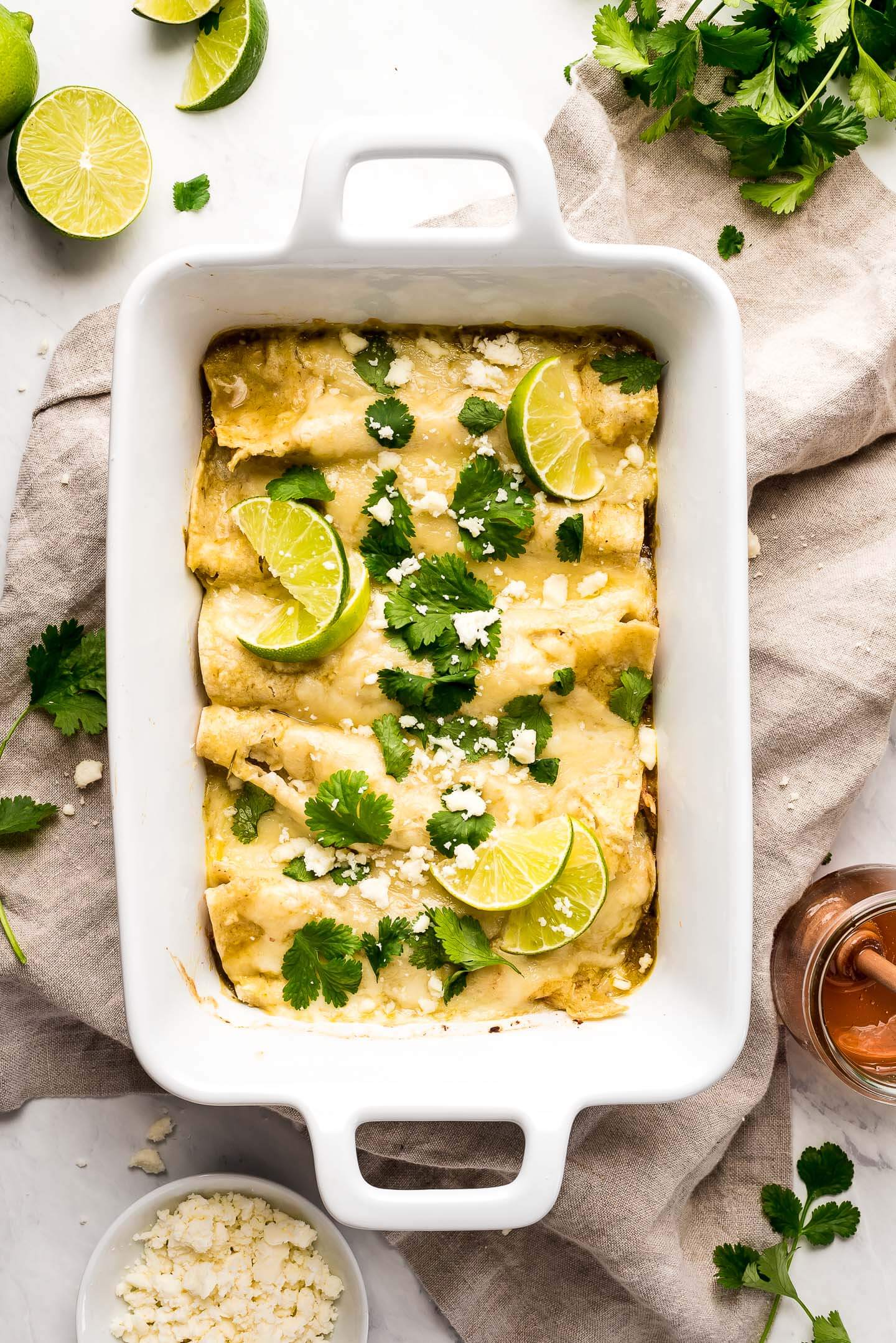 A white baking dish with Honey Lime Chicken Enchiladas topped with cilantro, lime wedges, and queso fresco and a jar of honey to the side.