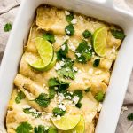 A white baking dish with green enchiladas topped with cilantro, lime wedges, and queso fresco.