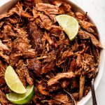 Pork Carnitas in a large white bowl garnished with lime wedges.