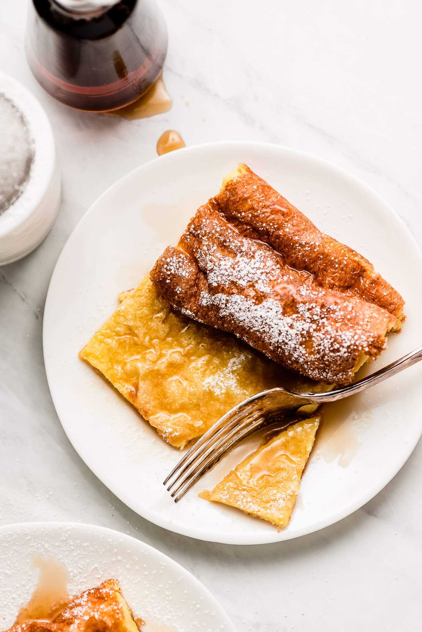 A fork cutting into in a piece of German Pancake covered in syrup and powdered sugar.