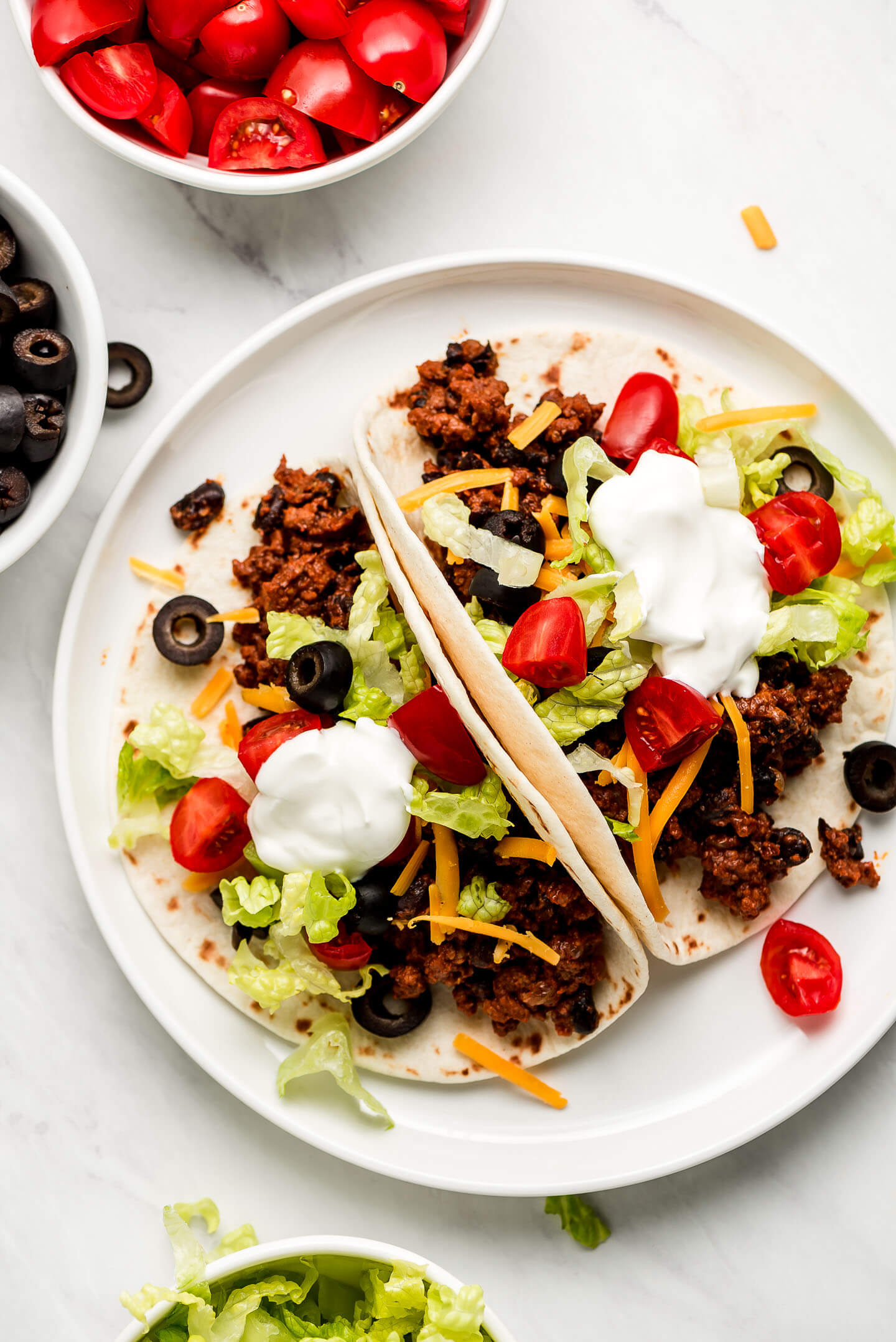 Two tortillas filled with ground beef filling and topped with lettuce, tomatoes, black olives, cheese, and sour cream.