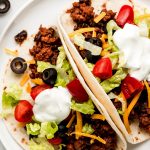 Two tortillas filled with ground beef filling and topped with lettuce, tomatoes, black olives, cheese, and sour cream.