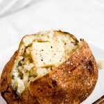 A plain Baked Potato on a plate cut open, fluffed and topped with a pat of butter, and sprinkled with pepper.