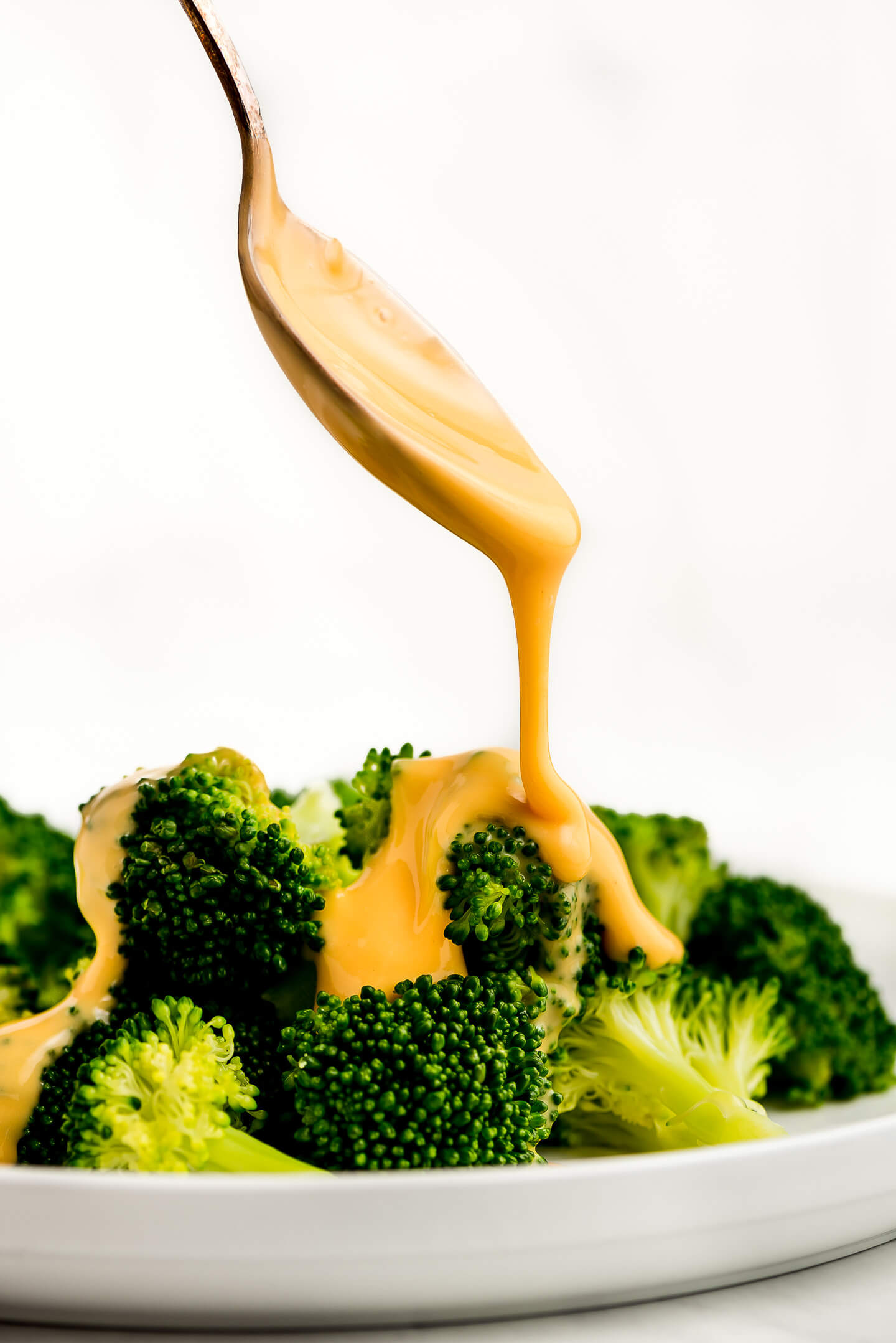 A spoon drizzling cheese sauce over steamed broccoli.