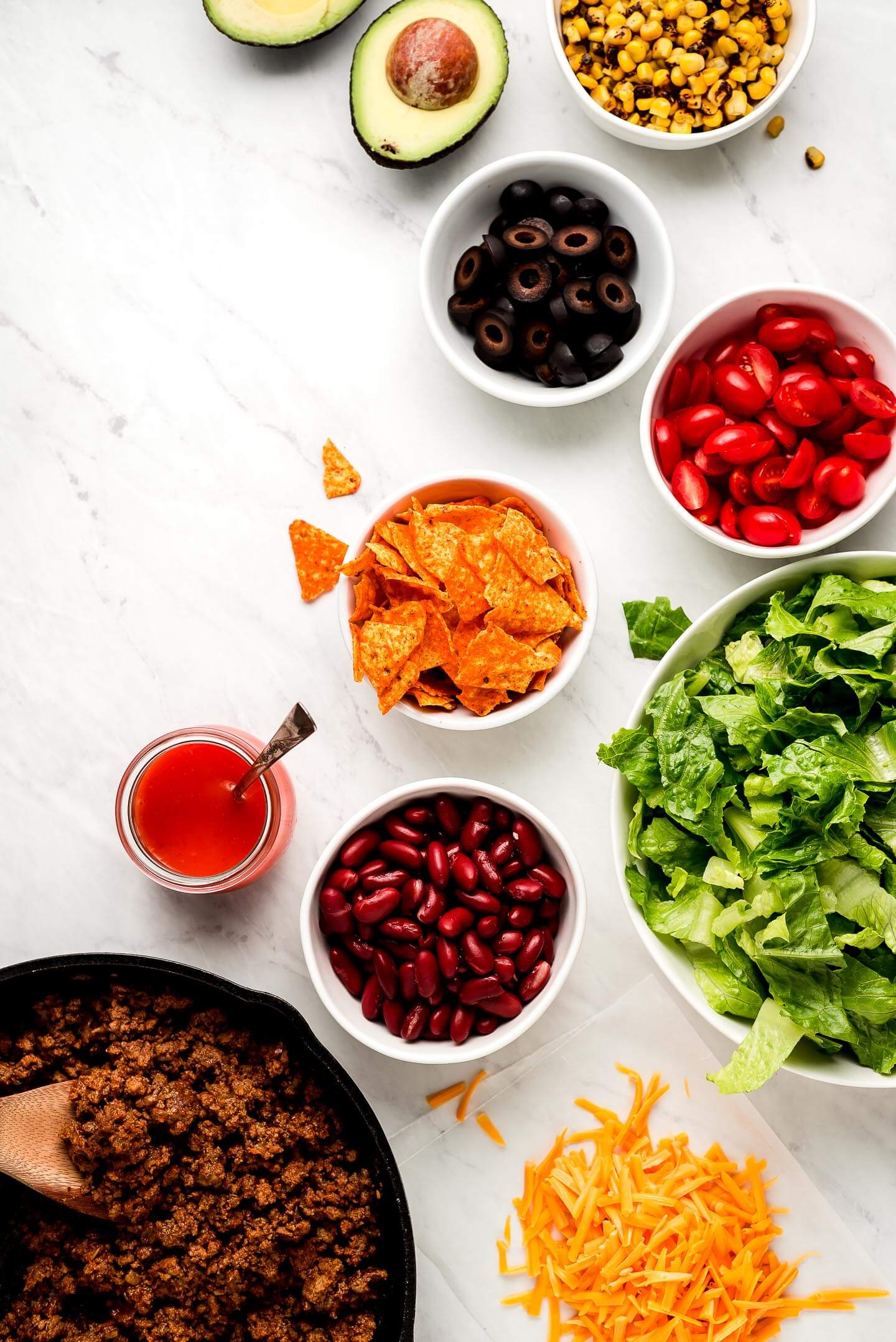 Ingredients on a marble surface- roasted corn, avocado, black olives, tomatoes, Doritos, lettuce, kidney beans, shredded cheese, taco meat, and dressing.