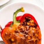 A red Stuffed Bell Pepper on a plate topped with cheese and a yellow stuffed pepper on another plate to the side.
