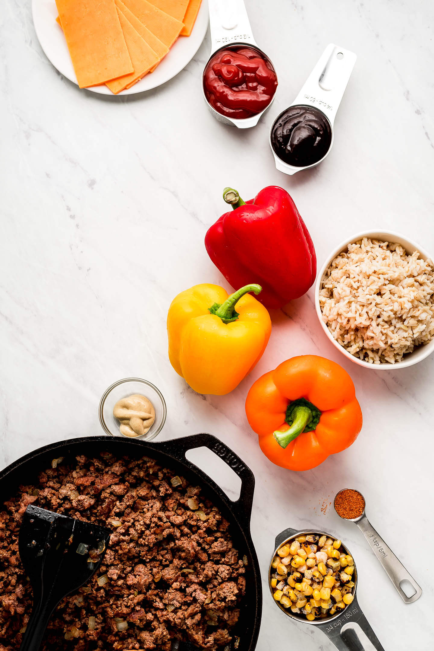 Ingredients on a marble surface- cheese, ketchup, barbecue sauce, bell peppers, rice, browned ground beef, corn, mustard, and seasoning.