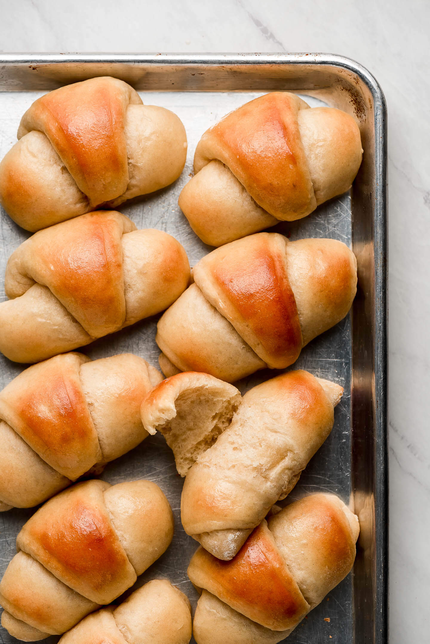 Homemade buttered Dinner Rolls lined up on a baking sheet with one pulled open to show fluffy interior.
