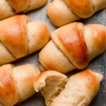 Homemade buttered Dinner Rolls lined up on a baking sheet with one pulled open to show fluffy interior.