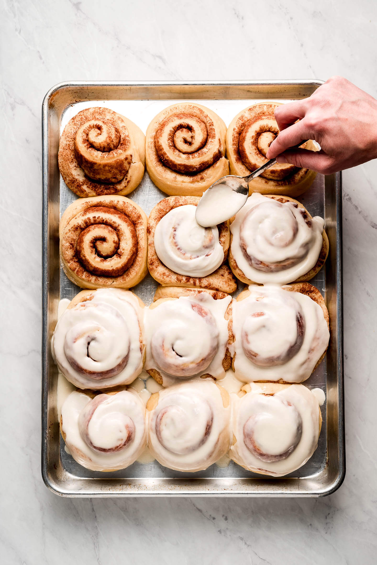 Spreading icing over cinnamon rolls on a sheet pan.