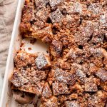 French Toast Bake on a marble surface with syrup, powdered sugar, and a piece of the casserole to the sides.