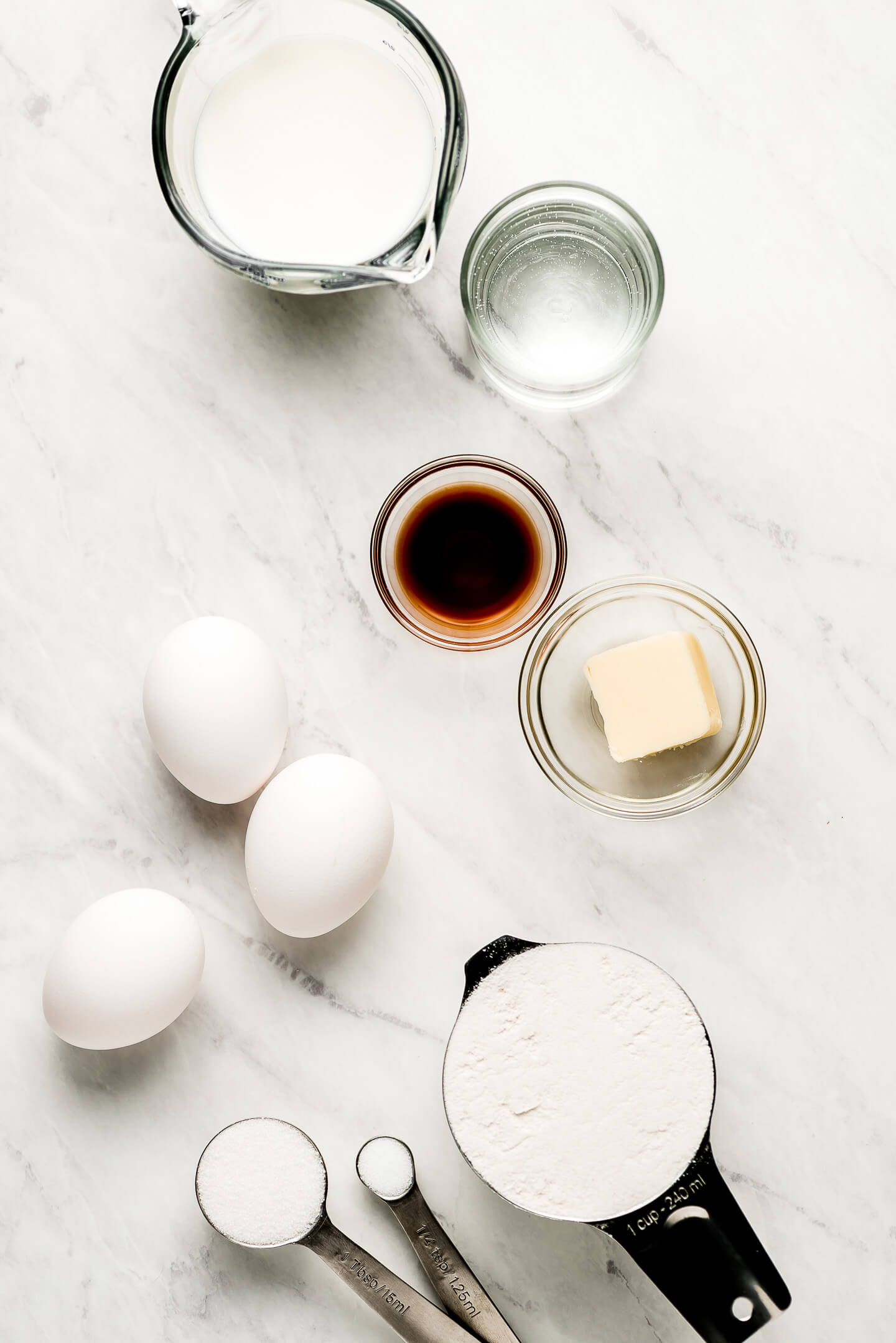Ingredients on a marble surface- milk, water, vanilla, butter, flour, eggs, and sugar.