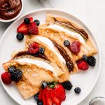 3 French Crepes on a plate stuffed with whipped cream, Nutella, and fresh fruit.