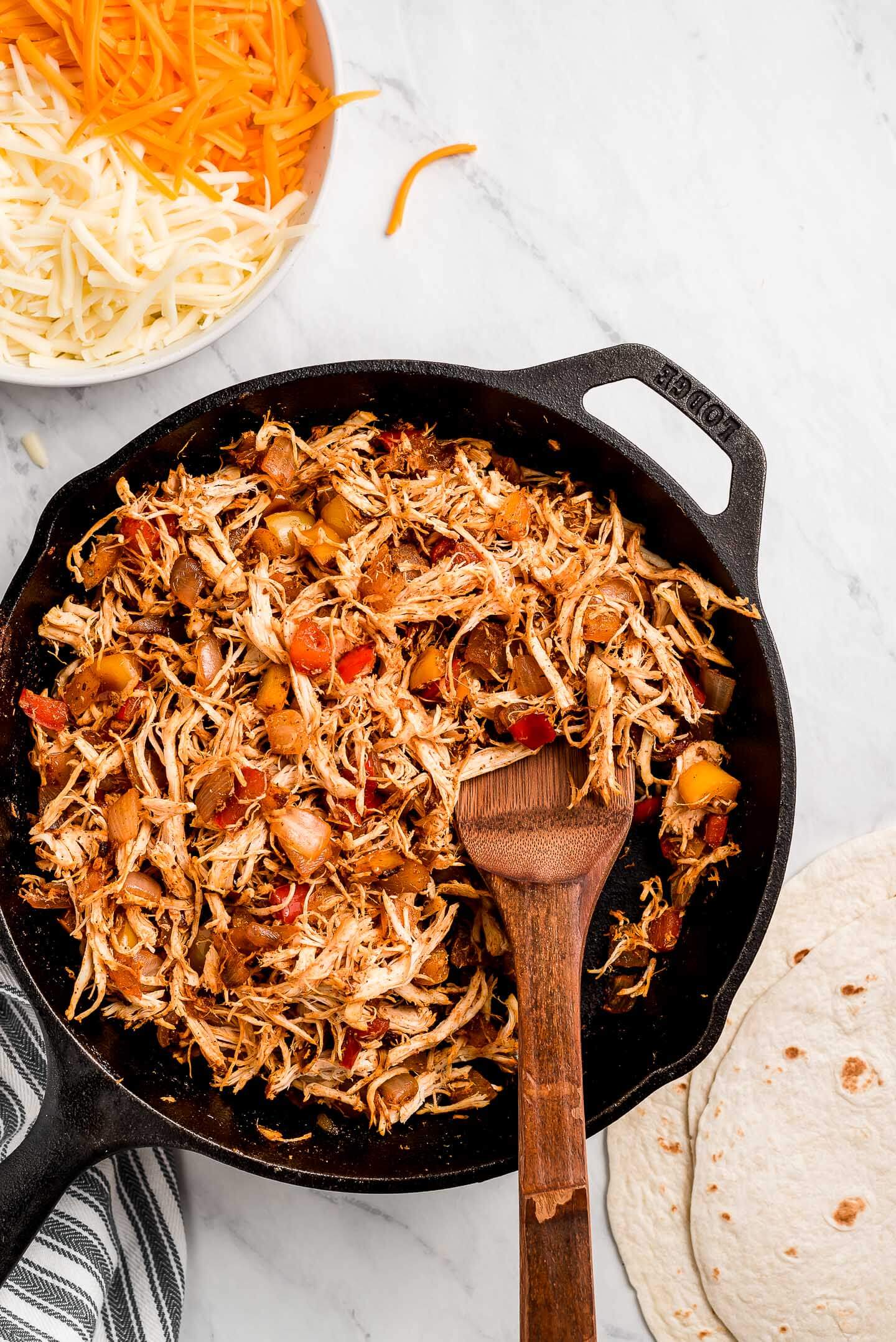 A skillet with chicken and peppers with a bowl of shredded cheese to the side.