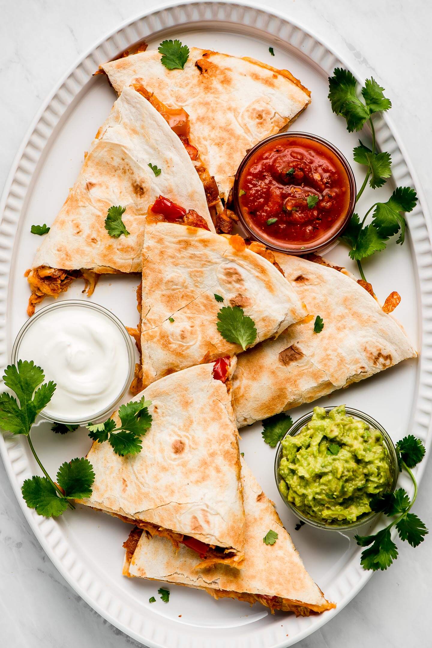 Triangles of quesadillas on a large platter with salsa, sour cream, guacamole, and cilantro.