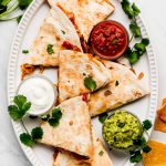 Triangles of quesadillas on a large platter with salsa, sour cream, and guacamole.