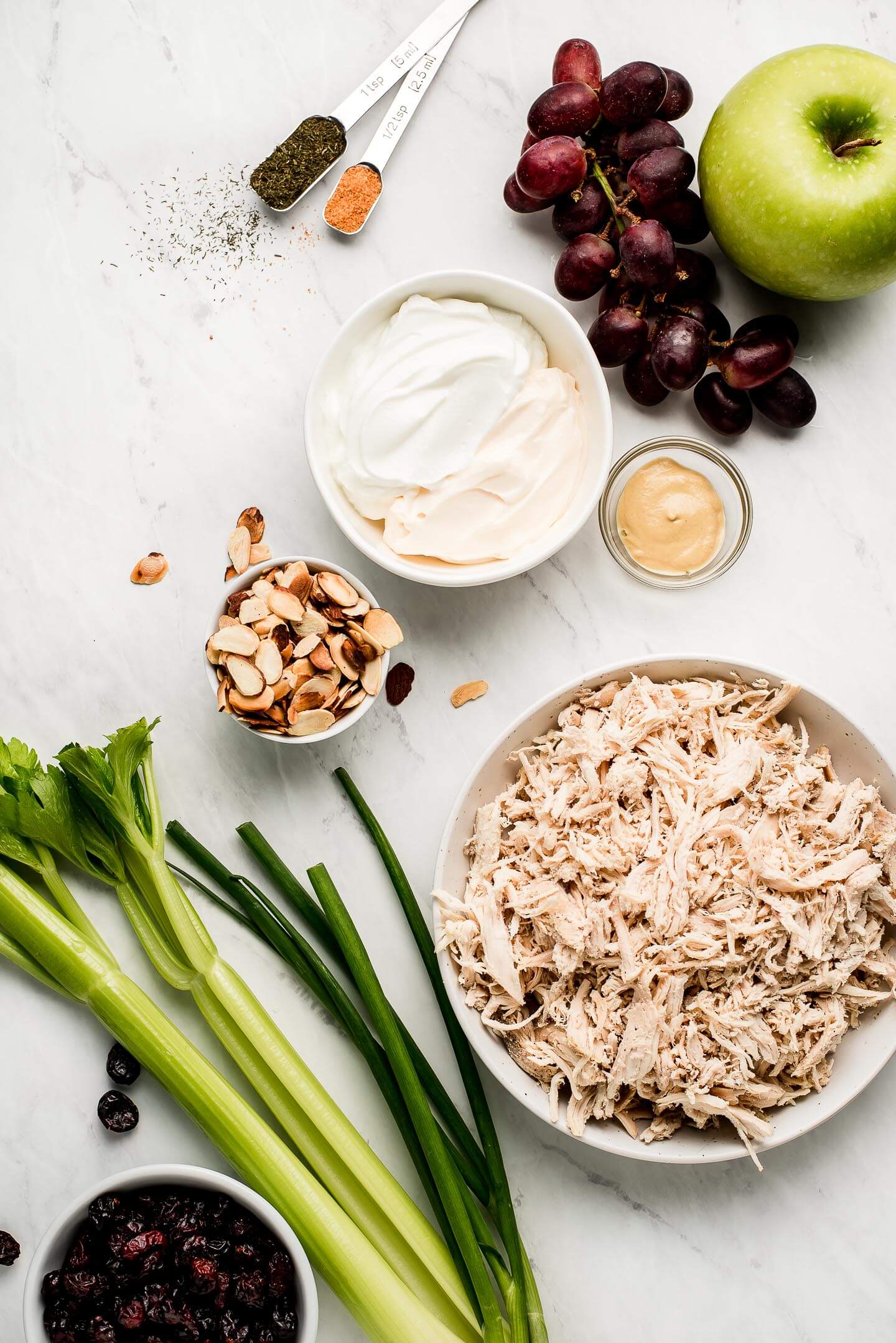 Ingredients on a marble surface- apple, grapes, seasoning, mayonnaise, nuts, mustard, shredded chicken, celery, onions, and Craisins.