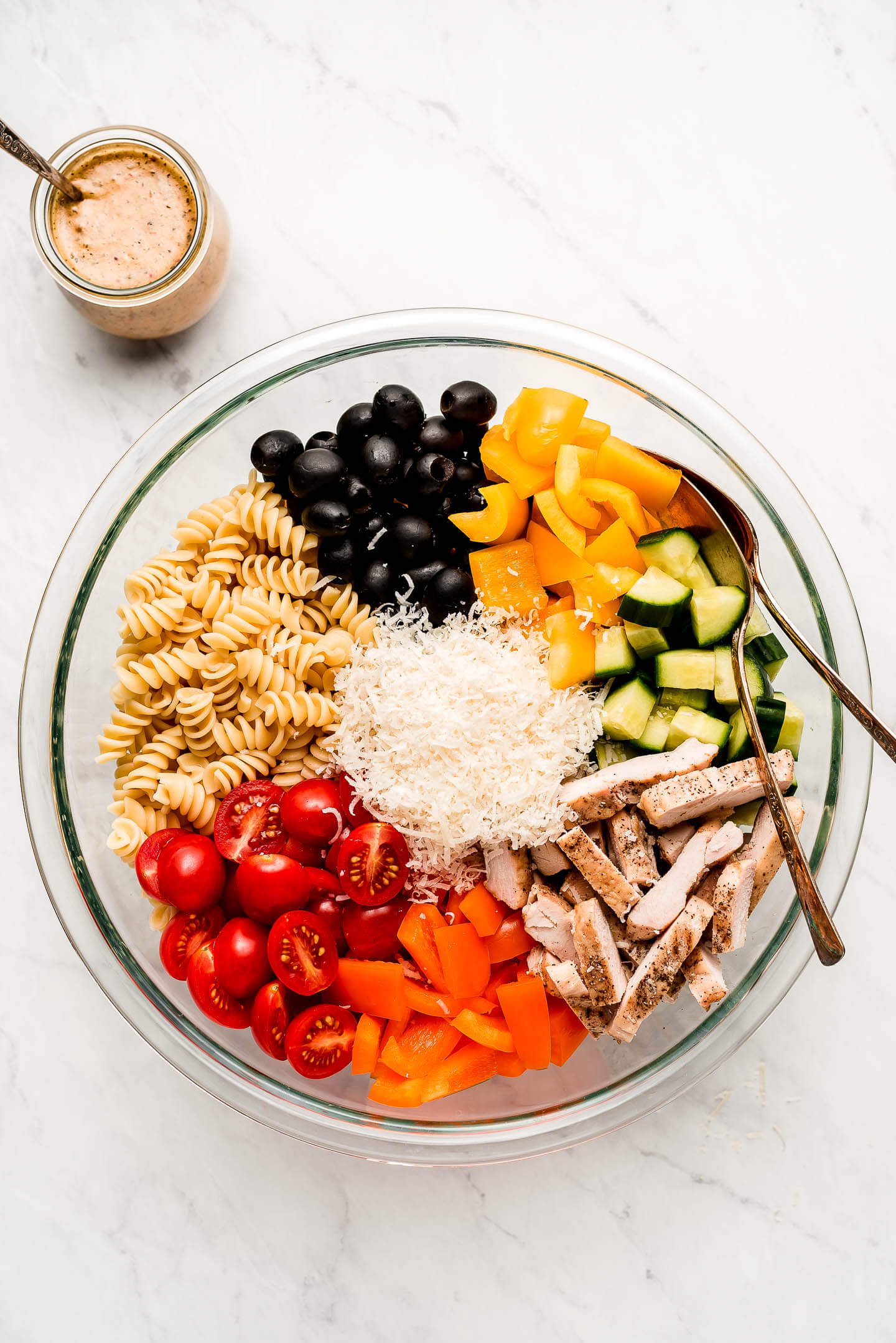 A bowl of separated ingredients- olives, peppers, cucumbers, grilled chicken, tomatoes, pasta, with a jar of dressing.