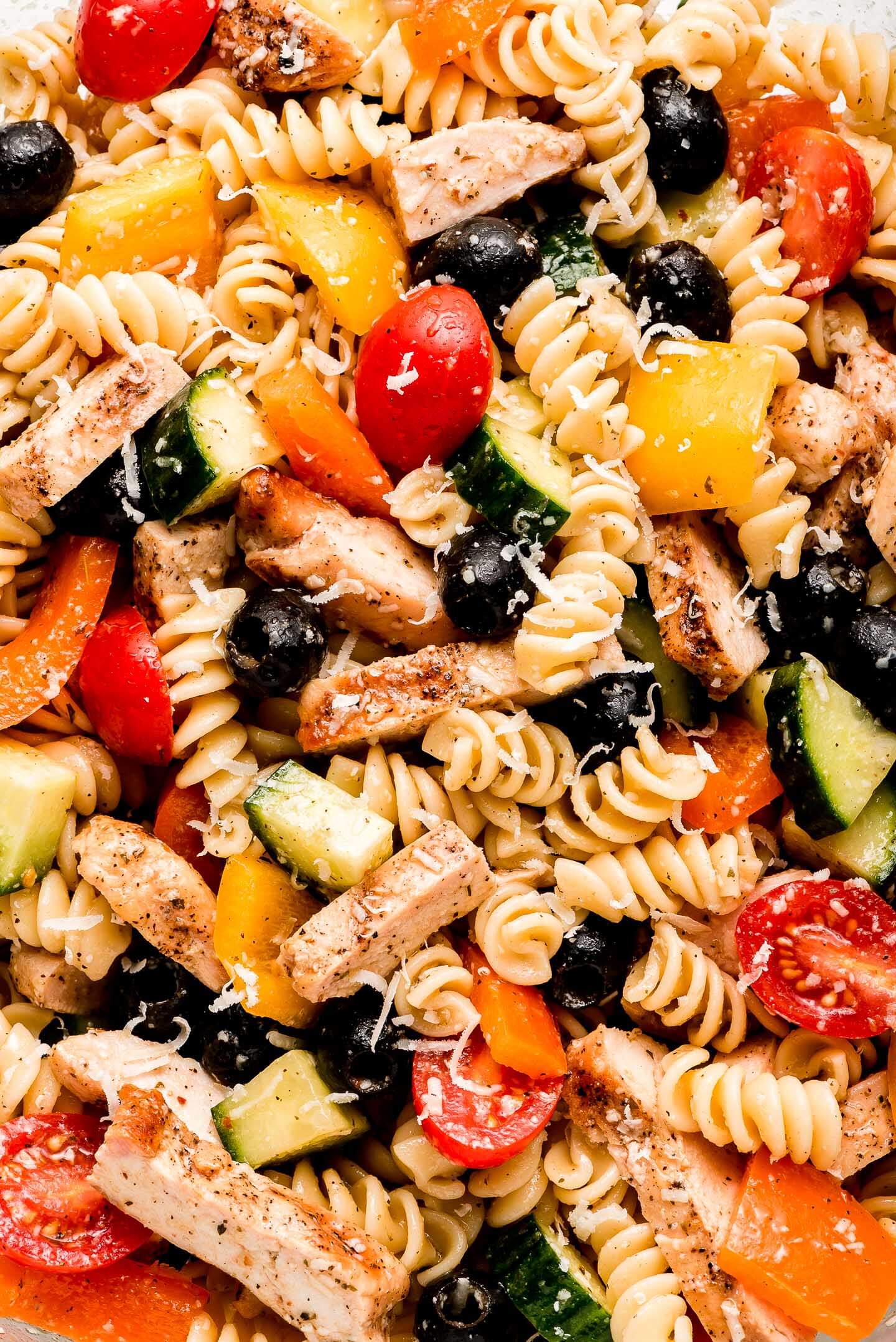 A close up shot of rotini pasta, sliced grilled chicken, black olives, grape tomatoes, cucumbers, and peppers.