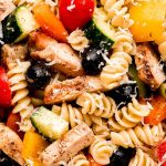 A close up shot of rotini pasta, sliced grilled chicken, black olives, grape tomatoes, cucumbers, and peppers.
