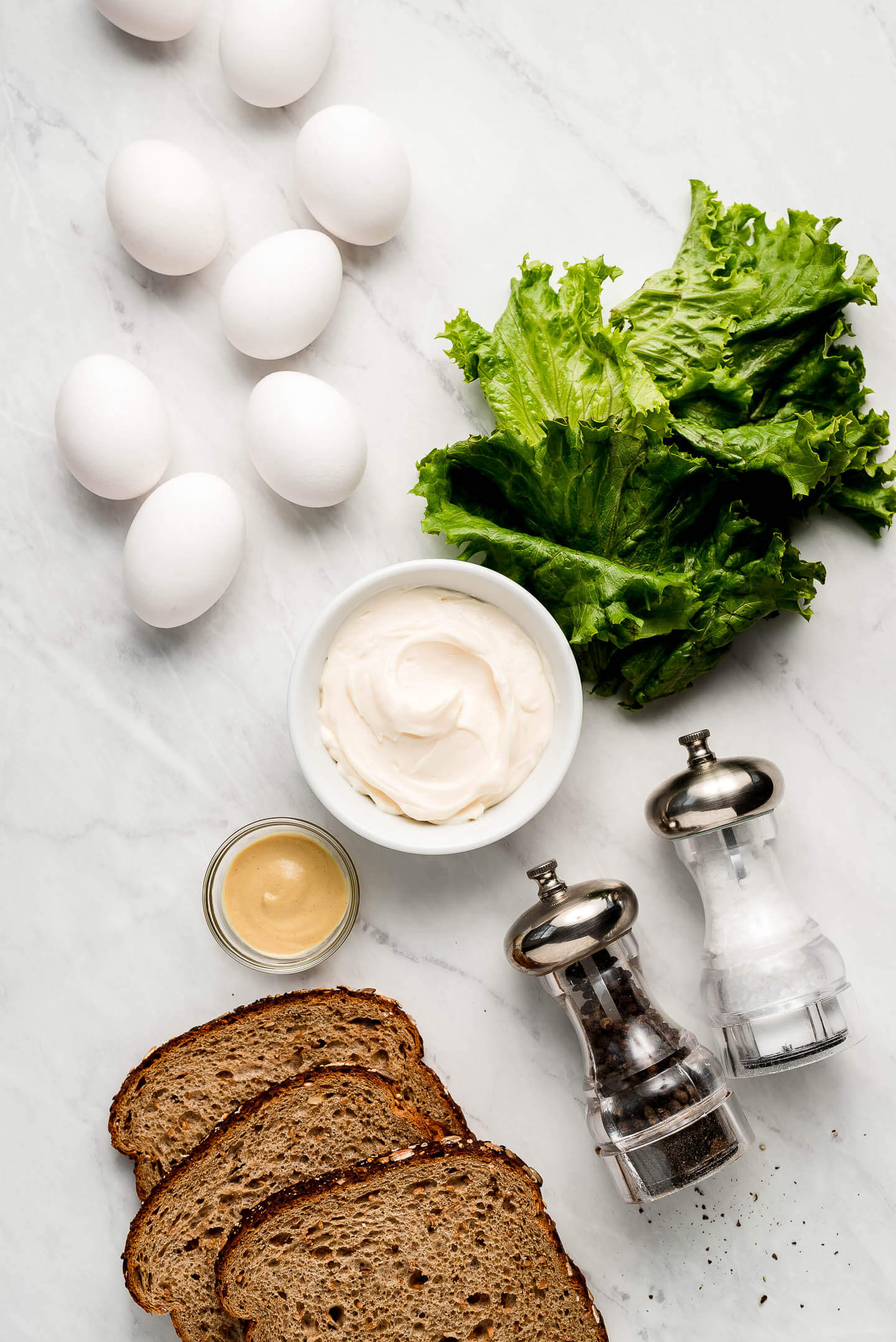 Ingredients on a marble surface- eggs, lettuce, mayo, mustard, bread, and salt & pepper.