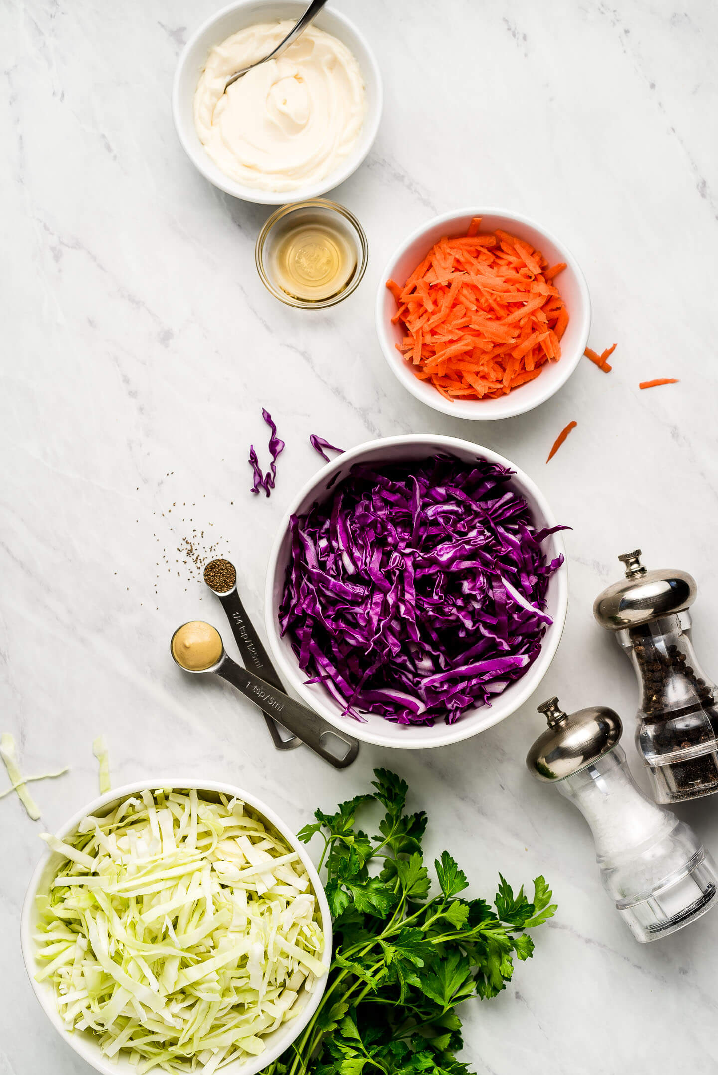 Ingredients on a marble surface- mayonnaise, vinegar, shredded carrots, red and green cabbage, and parsley.