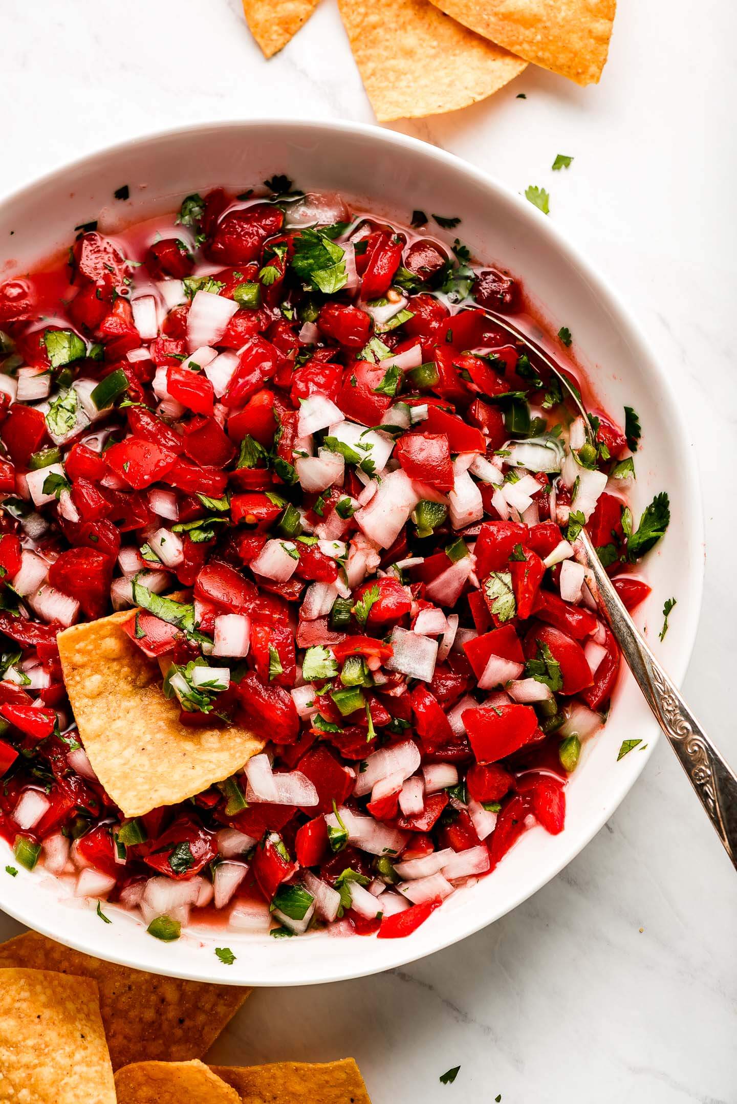 Pico de Gallo in a large white bowl with a spoon on the side and chips around the bowl.