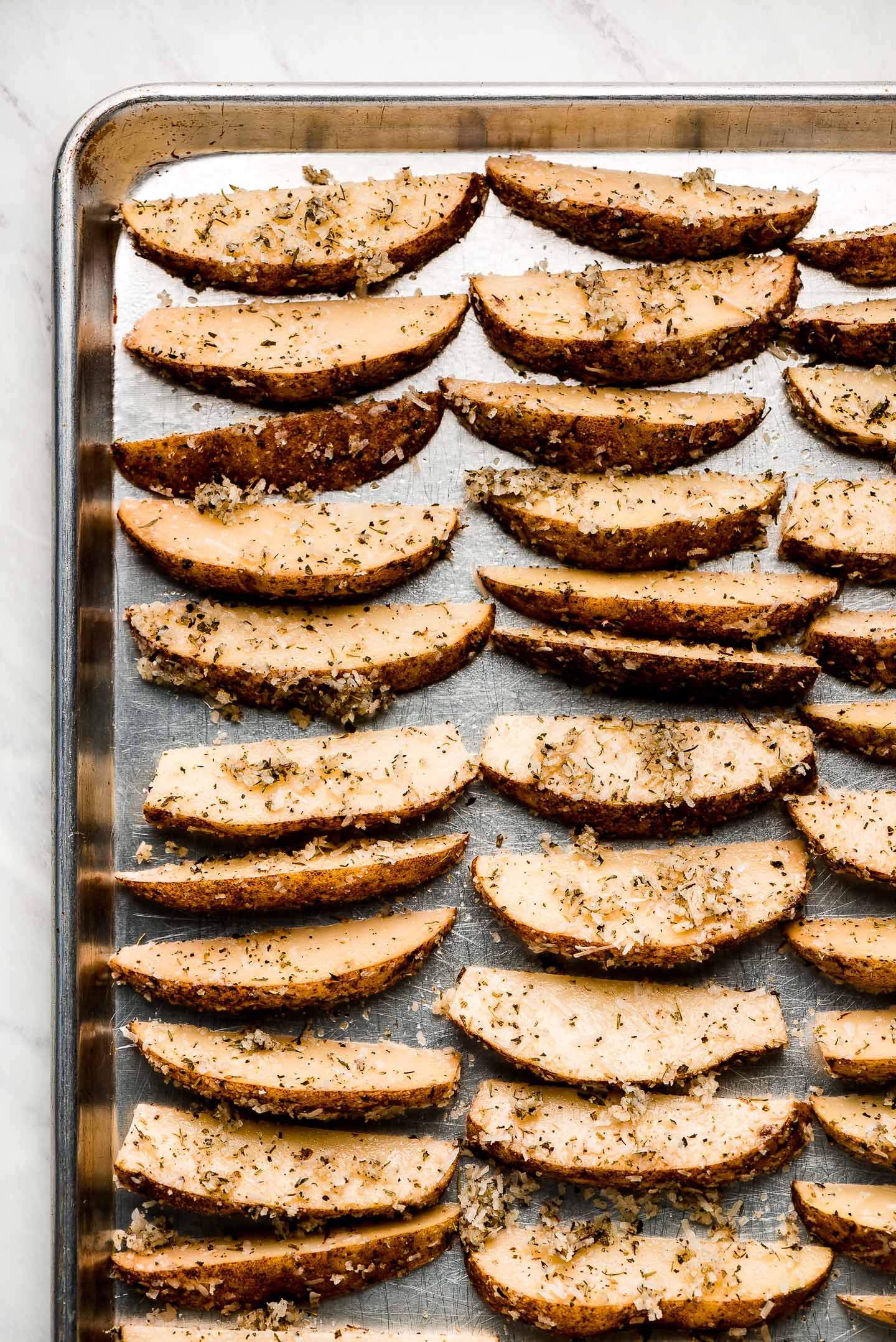 Raw potato wedges with herbs and parmesan on a baking sheet.