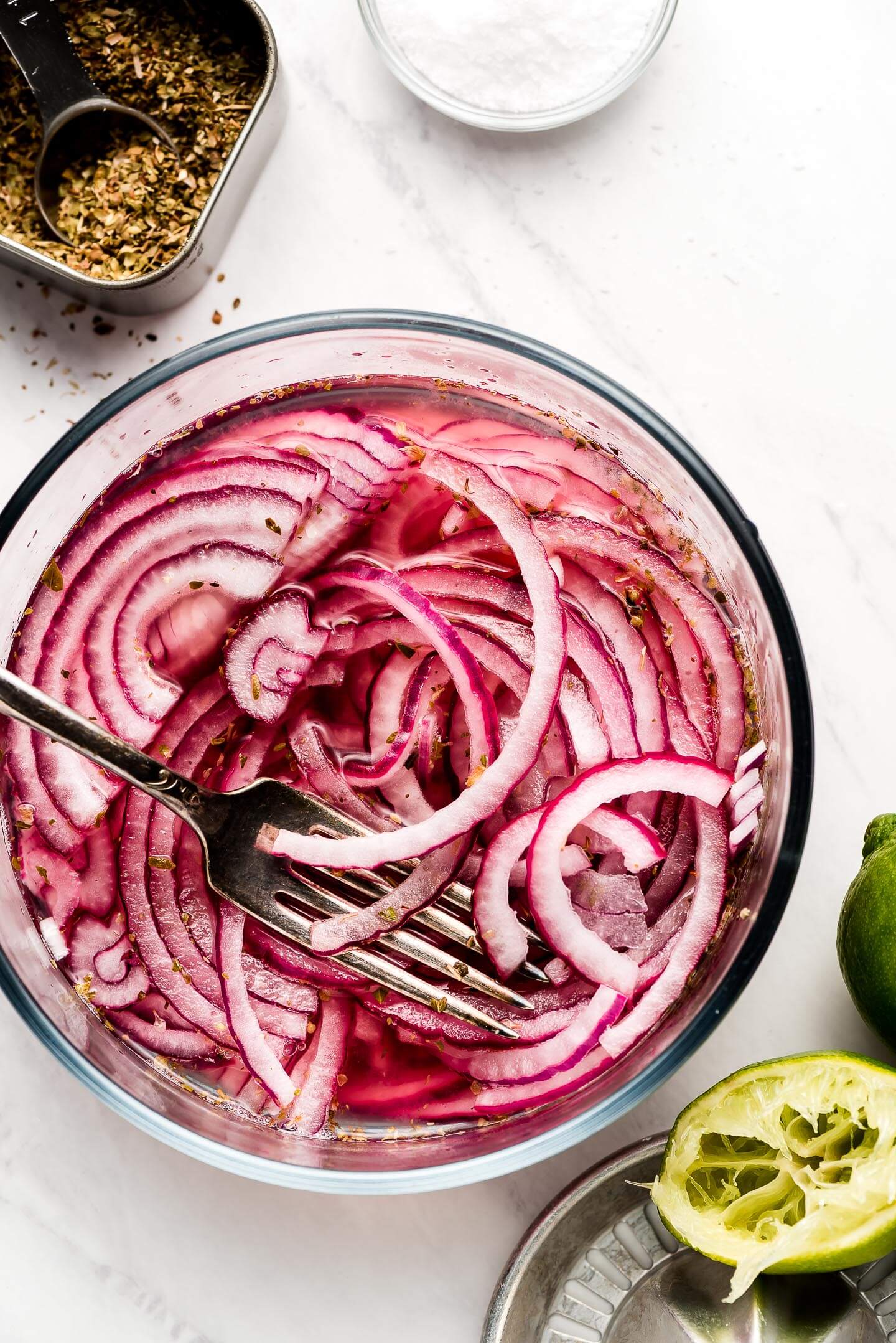Glass bowl with Mexican Pickled Onions with containers of oregano, salt, and limes around.