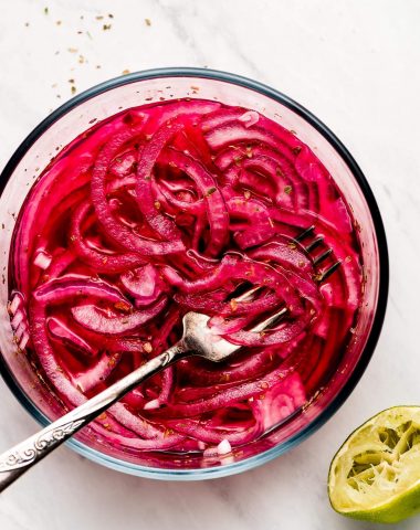 A glass bowl with marinated pink Mexican Pickled Onions with a squeezed lime to the side.