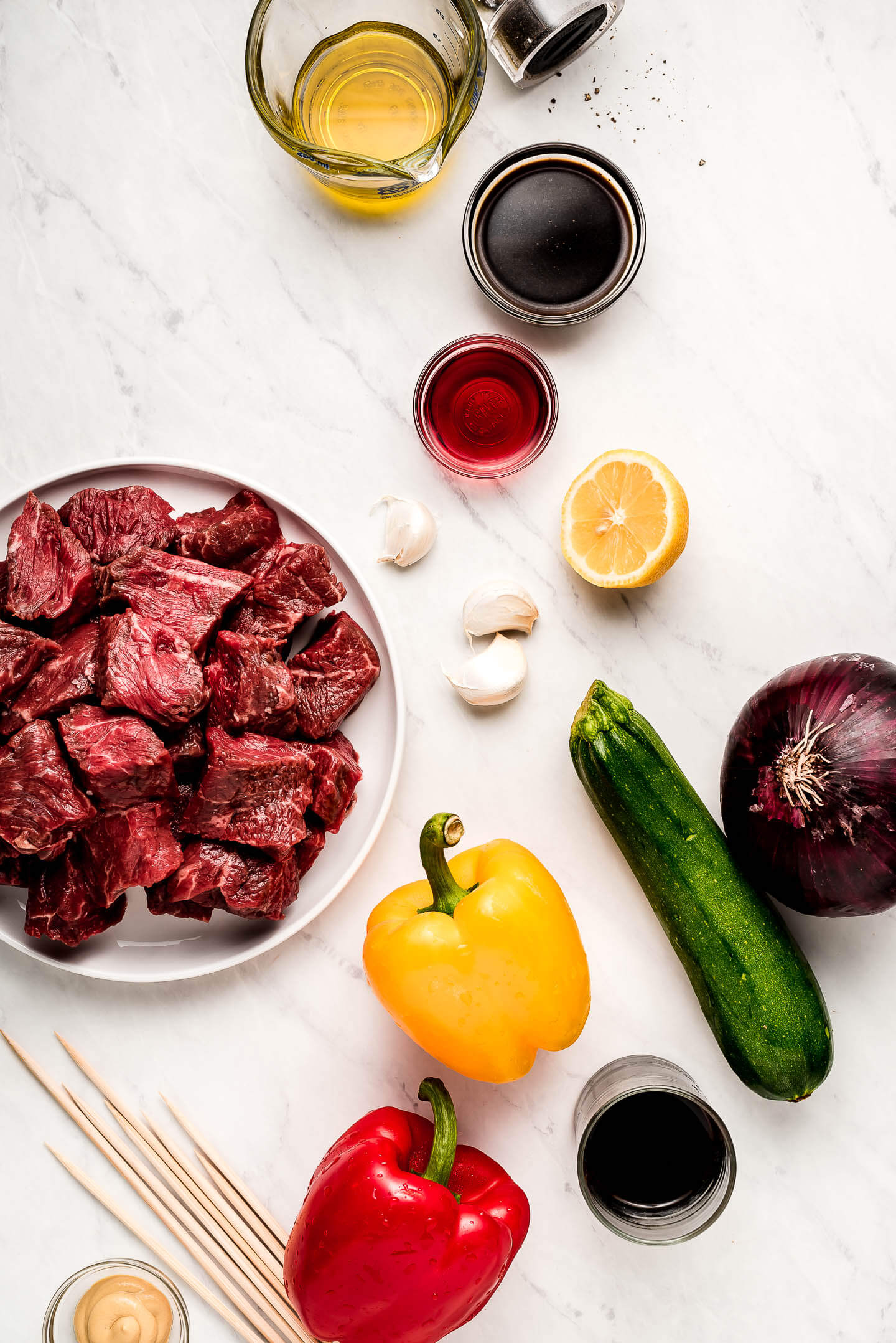 Ingredients on a marble surface- oil, soy sauce, vinegar, lemon, beef, peppers, zucchini, onion, and skewers.