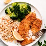 A plate of broccoli, rice, lemon wedge, and breaded tilapia that is flaked with a fork.
